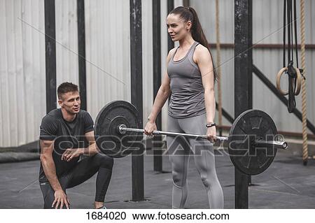 Stock Image - Male instructor looking at young woman crossfit training at gym. Fotosearch