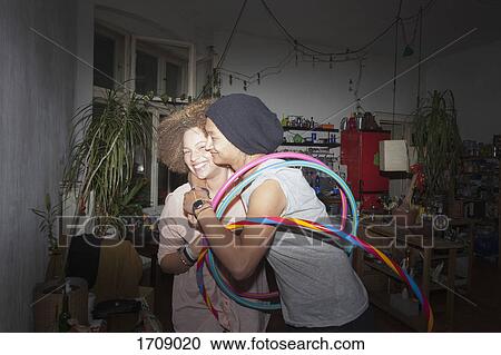 Stock Image - Man embracing smiling girlfriend while holding plastic hoops at home. Fotosearch