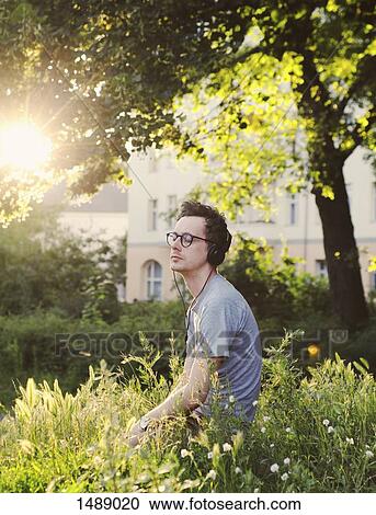 Stock Image - Man enjoying music through headset while sitting in park. Fotosearch