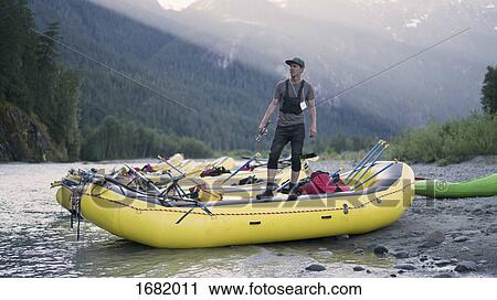 Stock Image - Man looking away while standing on raft at riverbank against mountain. Fotosearch