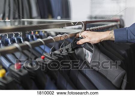 Stock Image - Man reaching for suit on hanger in menswear shop. Fotosearch