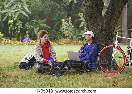 Man with laptop talking to friend while sitting on grassy field at park View Large Photo Image Stock Photo - Man with laptop talking to friend while sitting on grassy field at park. Fotosearch