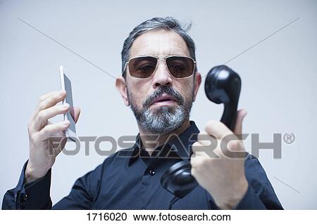 Stock Image - Mature businessman holding telephone receiver and mobile phone against white background. Fotosearch