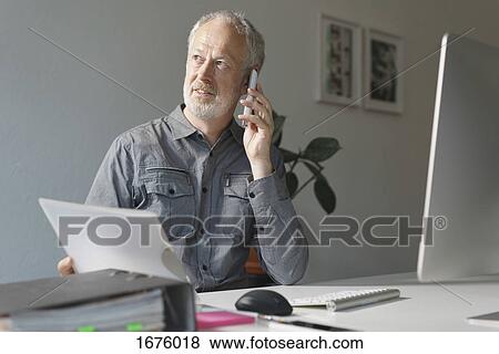 Stock Photo - Mature businessman using mobile phone while sitting at desk in office. Fotosearch