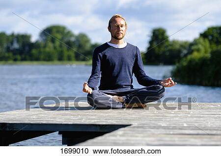 Stock Image - Mature man practicing lotus position on pier by lake against sky. Fotosearch