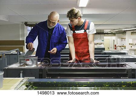 Stock Image - Mature worker showing colleague ink container on printer at printing press. Fotosearch