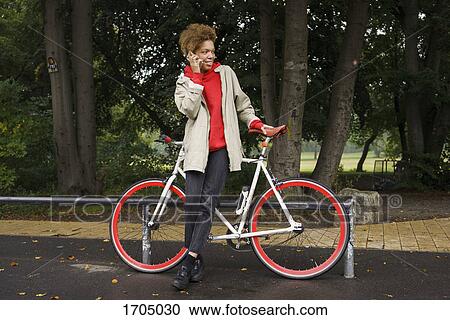 Mid adult woman talking on mobile phone while standing with bicycle at park View Large Photo Image Stock Image - Mid adult woman talking on mobile phone while standing with bicycle at park. Fotosearch