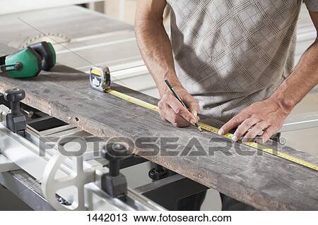 Stock Image - Midsection of male carpenter measuring plank on sliding table saw in workshop. Fotosearch