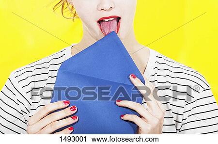 Stock Image - Midsection of woman licking blue envelope against yellow background. Fotosearch