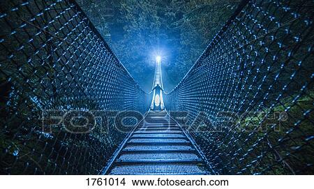 Picture - Mysterious silhouetted person on suspension bridge in woods at night. Fotosearch