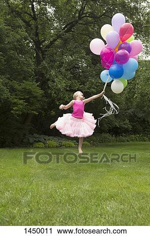 Stock Image - Playful girl jumping while holding balloons at backyard. Fotosearch
