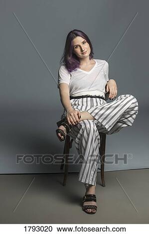 Stock Image - Portrait confident and fashionable young woman sitting on chair against gray background. Fotosearch