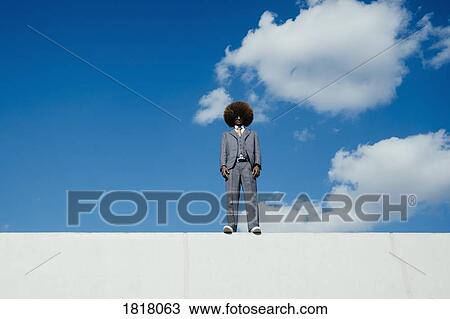 Stock Image - Portrait confident, cool, well-dressed young man with afro standing on wall against sky. Fotosearch