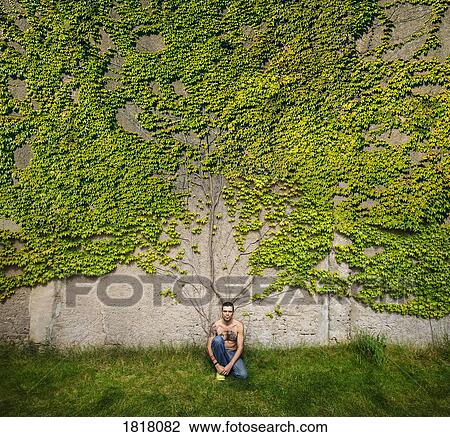 Stock Image - Portrait man with tattooed chest sitting at wall overgrown with ivy. Fotosearch