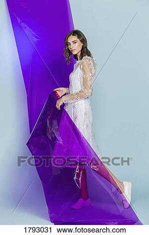 Stock Image - Portrait of a female fashion model posing with purple plastic sheet against blue background. Fotosearch
