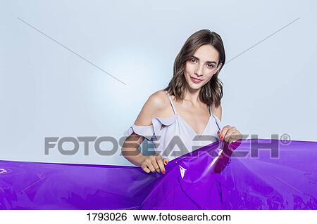 Stock Photograph - Portrait of a female fashion model posing with purple plastic sheet against blue background. Fotosearch