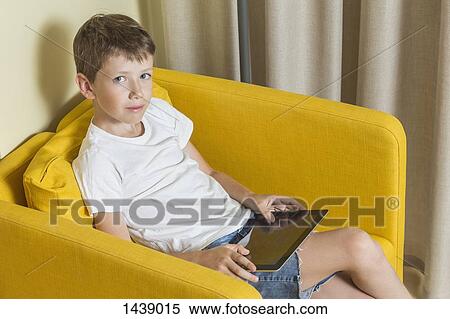 Stock Photography - Portrait of boy holding digital tablet while sitting on armchair at home. Fotosearch