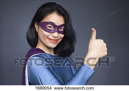 Picture - Portrait of confident female superhero gesturing thumbs up against gray background. Fotosearch