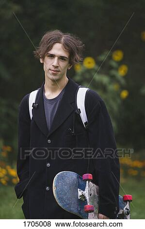 Stock Photo - Portrait of confident young man carrying skateboard while standing at park. Fotosearch