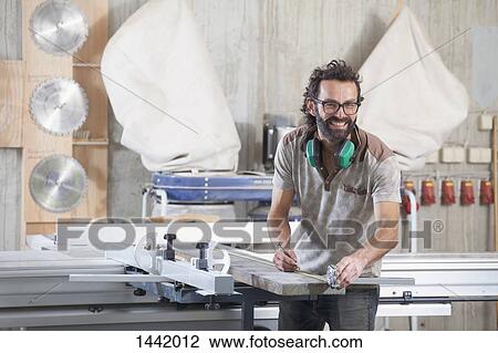 Stock Image - Portrait of happy male carpenter measuring lumber in workshop. Fotosearch