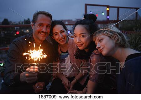 Stock Photo - Portrait of happy man holding sparkler while sitting with female friends at night. Fotosearch