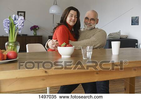 Portrait of happy mature couple embracing and sitting by wooden table at home View Large Photo Image Stock Photography - Portrait of happy mature couple embracing and sitting by wooden table at home. Fotosearch
