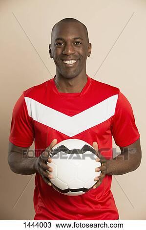 Stock Image - Portrait of happy soccer player holding ball against colored background. Fotosearch