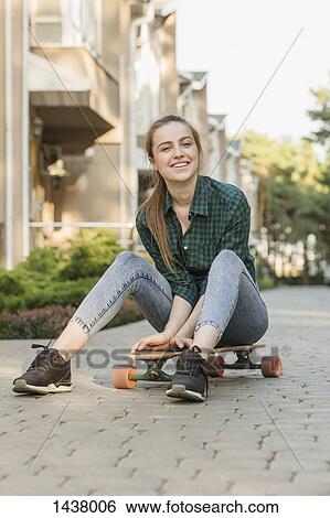 Stock Photograph - Portrait of happy young woman sitting on skateboard outdoors. Fotosearch