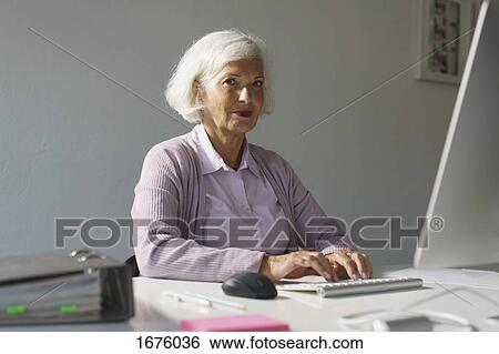 Stock Photograph - Portrait of senior woman using computer while sitting against wall in office. Fotosearch