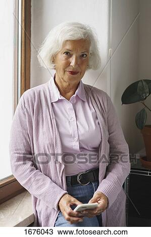 Stock Image - Portrait of senior woman with mobile phone leaning on window sill at office. Fotosearch