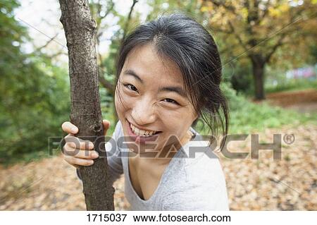Stock Photo - Portrait of smiling woman holding tree trunk while standing at park. Fotosearch