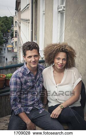 Stock Photo - Portrait of smiling young friends sitting by wall in balcony. Fotosearch