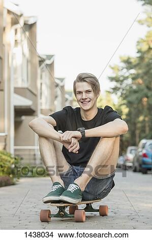 Picture - Portrait of smiling young man sitting on skateboard outdoors. Fotosearch