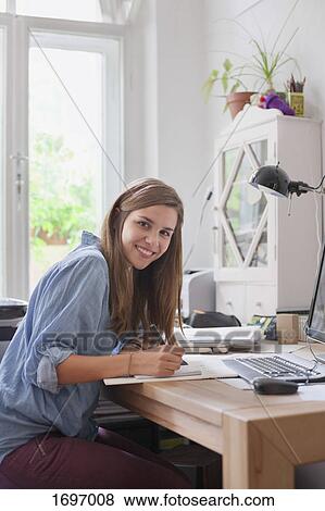 Portrait of smiling young woman writing at computer table in room View Large Photo Image Stock Photo - Portrait of smiling young woman writing at computer table in room. Fotosearch