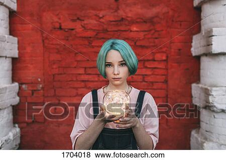 Stock Photo - Portrait of teenage girl holding illuminated string lights while standing against brick wall. Fotosearch