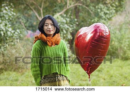 Picture - Portrait of woman by red heart shape balloon while standing at park. Fotosearch