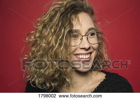 Stock Image - Portrait of woman smiling with curly hair and wearing glasses against red background. Fotosearch