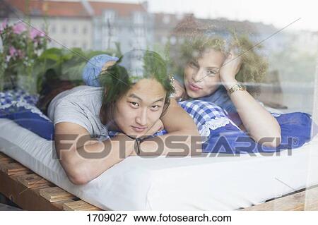 Stock Photo - Portrait of young couple lying on bed seen through glass window at home. Fotosearch