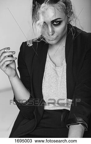 Stock Image - Portrait of young woman smoking cigarette while sitting against gray background. Fotosearch