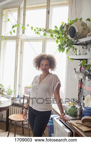 Stock Image - Portrait of young woman standing by kitchen counter against window at home. Fotosearch