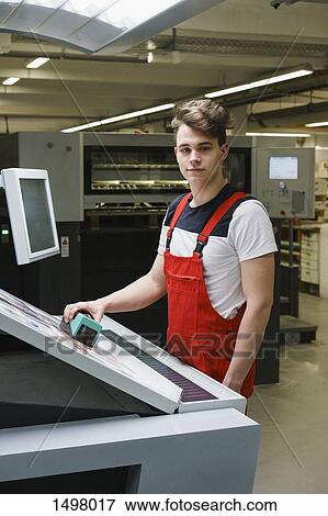 Stock Photo - Portrait of young worker holding scanner over printout with machinery at printing press. Fotosearch