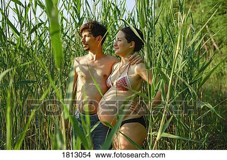 Picture - Pregnant couple in bikini and swim trunks standing in rural green corn field. Fotosearch