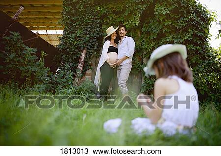 Stock Image - Pregnant couple watching daughter sitting in grass. Fotosearch