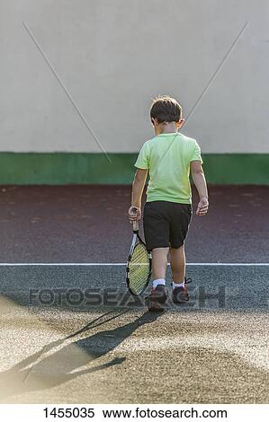 Stock Photography - Rear view of boy holding tennis racket and walking at court. Fotosearch