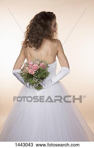 Stock Image - Rear view of bride holding bouquet against colored background. Fotosearch
