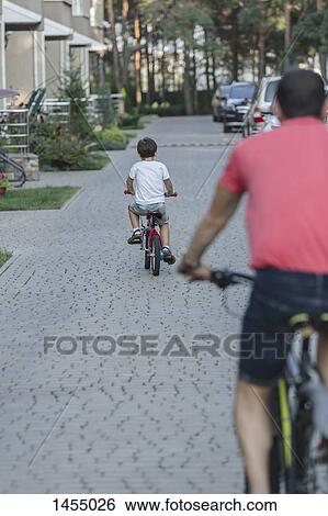Stock Photograph - Rear view of father and son riding bicycle on street. Fotosearch