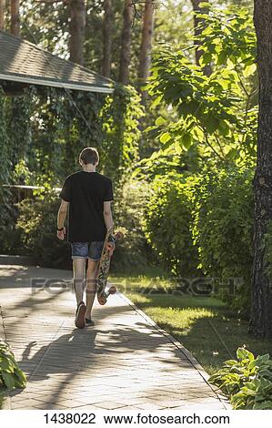 Rear view of man carrying skateboard while walking on footpath at park View Large Photo Image Stock Image - Rear view of man carrying skateboard while walking on footpath at park. Fotosearch
