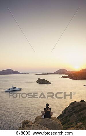 Rear view of woman sitting on rock formation while looking at sea against sky during sunset View Large Photo Image Stock Image - Rear view of woman sitting on rock formation while looking at sea against sky during sunset. Fotosearch