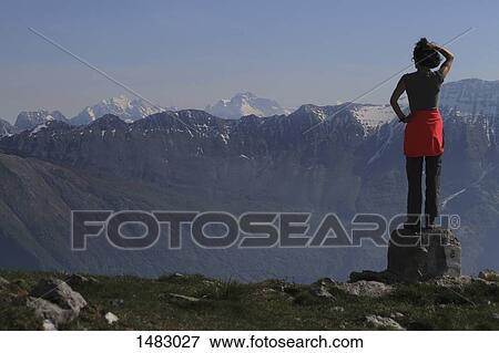Stock Photo - Rear view of woman standing on rock against mountains. Fotosearch