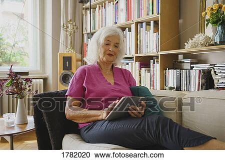Stock Image - Senior woman using digital tablet on living room sofa. Fotosearch